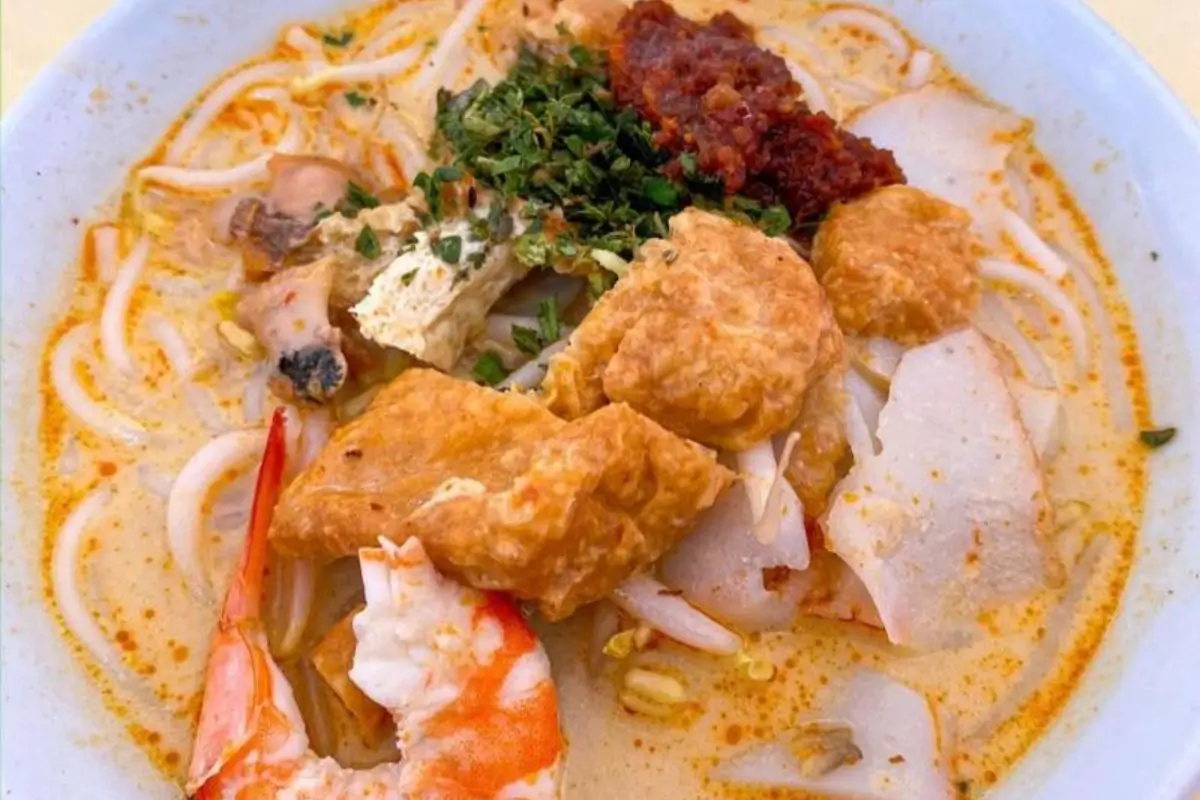 A bowl of Singapore Laksa, a popular spicy noodle soup, made with a creamy coconut milk-based broth rich in chili and spices. It is loaded with ingredients including rice noodles, shrimp, fish cakes, tofu, and bean sprouts, and is garnished with fresh cilantro, chili paste, and a squeeze of fresh lime