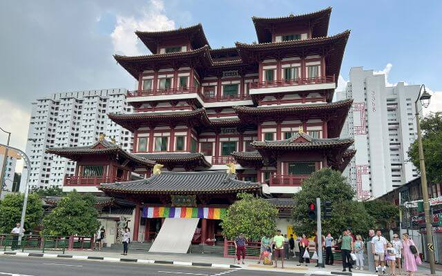 The Buddha Tooth Relic Temple in Singapore, a striking multi-tiered red and gold traditional structure, stands prominently against the cityscape, with visitors waiting to cross the street in front.