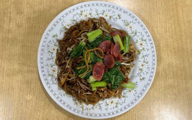 A plate of Char Kway Teow, a traditional stir-fried flat rice noodle dish, with slices of Chinese sausage, bean sprouts, and green vegetables, served on a decorative floral-patterned plate.