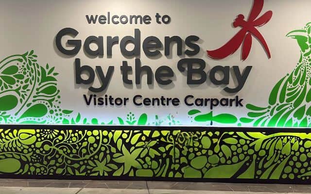 Signboard at the entrance of Gardens by the Bay in Singapore, featuring bold lettering and a red dragonfly graphic above a green foliage design, indicating the Visitor Centre Carpark.