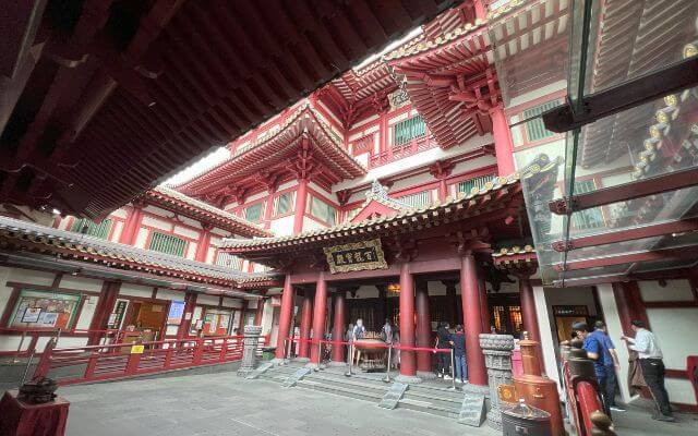 Inside the Buddha Tooth Relic Temple in Singapore, with a view of the grand red and gold entrance to the main prayer hall, intricate architectural details, and visitors moving about.