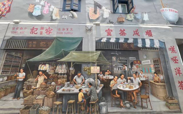 A street mural in Singapore's Chinatown vividly captures a scene from the past, featuring a 'Kopitiam' or traditional coffee shop and a provision shop, with local residents engaging in daily activities against a backdrop of hanging laundry.