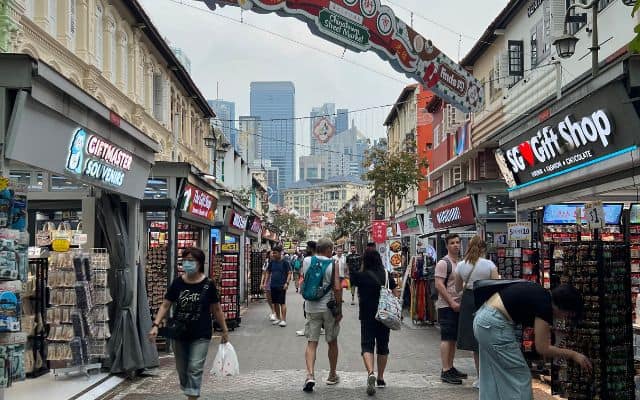 A lively street in Singapore's Chinatown, bustling with tourists and locals browsing through a variety of gift shops and stalls under colorful signage, set against a backdrop of historic shop houses and modern skyscrapers.