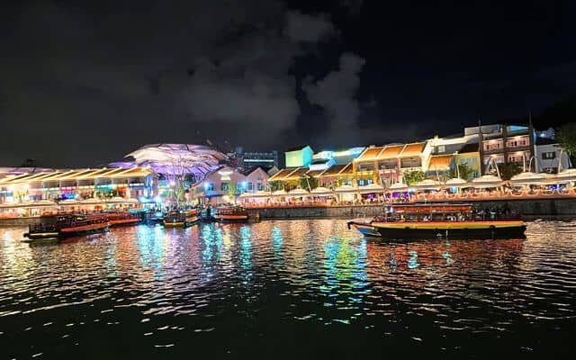 Nighttime view of Singapore's Clarke Quay, its vibrant illuminated facades reflecting on the calm river waters. Traditional bumboat cruises with passengers traverse the serene river, contributing to the lively atmosphere of this popular dining and entertainment hub.