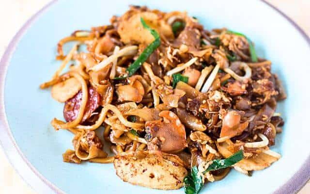 A delectable plate of Char Kway Teow, a classic Singaporean stir-fry noodle dish, complete with juicy cockles, bean sprouts, and Chinese sausage slices, served on a pale blue plate.