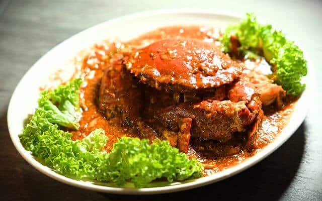 A savory Singaporean chili crab dish garnished with lettuce, presented on a white plate against a dark table backdrop, highlighting the rich red sauce coating the succulent crab.
