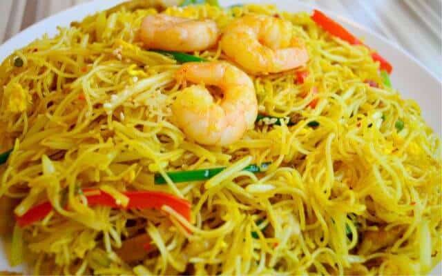 Close-up of Singapore-style fried rice noodles seasoned with curry, mixed with slices of red bell pepper, green onions, and topped with succulent shrimp, served on a white plate.