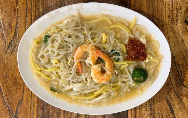 A plate of Singapore Hokkien noodles, featuring a mix of rice noodles and egg noodles, tossed with prawn, squid rings, and thin slices of pork, garnished with a spoonful of red sambal chili and lime on the side.
