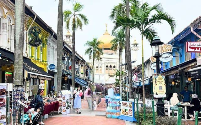 The cultural richness of Kampong Gelam in Singapore is on display with its colorful shop houses lining the street, leading to the golden dome of the Sultan Mosque in the background. People wander the lively market area, dotted with street stalls, under the tropical palm trees, contributing to the district's historical charm.
