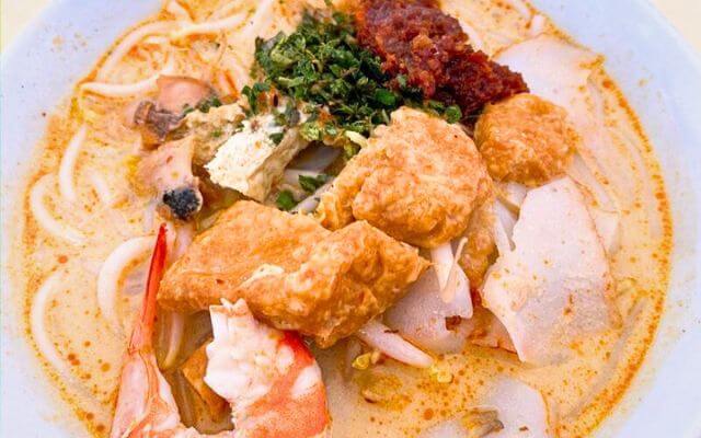 A bowl of Singaporean laksa, a spicy noodle soup, with prawns, tau pok (fried bean-curd puffs), sliced fish cakes, and cockles, garnished with chopped laksa leaves and a dollop of sambal chili paste