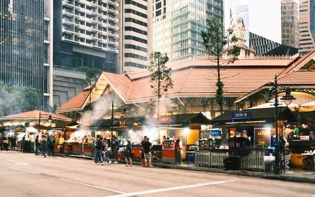 The bustling atmosphere of Lau Pa Sat food market in Singapore, with smoke wafting from street food stalls, people milling about, and the historical clock tower visible atop the structure, set against a backdrop of modern skyscrapers.