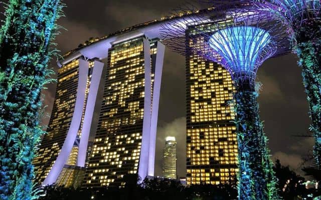 Night view of Marina Bay Sands in Singapore, seen from Gardens by the Bay with the futuristic Supertree Grove structures illuminated in blue, contrasting against the warmly lit hotel towers and the evening sky.