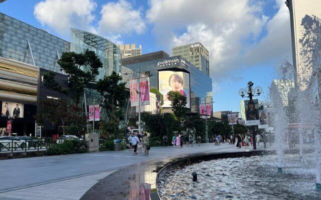 A vibrant scene on Orchard Road, Singapore's upscale shopping area, showcasing the grand Pullman hotel, large advertising screens, and lush street-side greenery. Pedestrians wander the promenade near glistening water features under a partly cloudy sky.