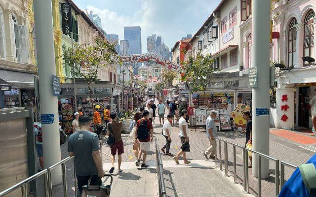 A bustling day on Pagoda Street in Singapore, as seen just after exiting from the MRT station. People walk among traditional shop houses with colorful facades under a clear blue sky, while modern skyscrapers loom in the background.