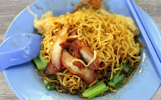 Plate of Singaporean Wantan Mee featuring yellow egg noodles topped with slices of char siu (barbecued pork), wantan dumplings, and green vegetables, served with a spoon on a blue plate.