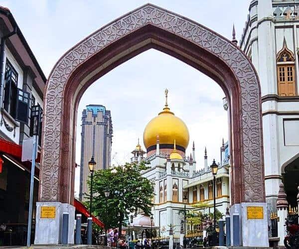 View through an ornate archway framing the massive golden dome of the Sultan Mosque in Singapore's Kampong Glam, capturing the essence of the historical and cultural landmark against a contrasting backdrop of modern high-rises.