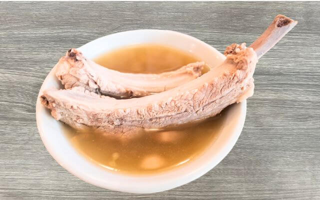 A bowl of Singaporean Bak Kut Teh featuring tender pork ribs immersed in a clear, garlicky broth. The pork ribs are large, with visible spices and garlic pieces enhancing the savory broth.