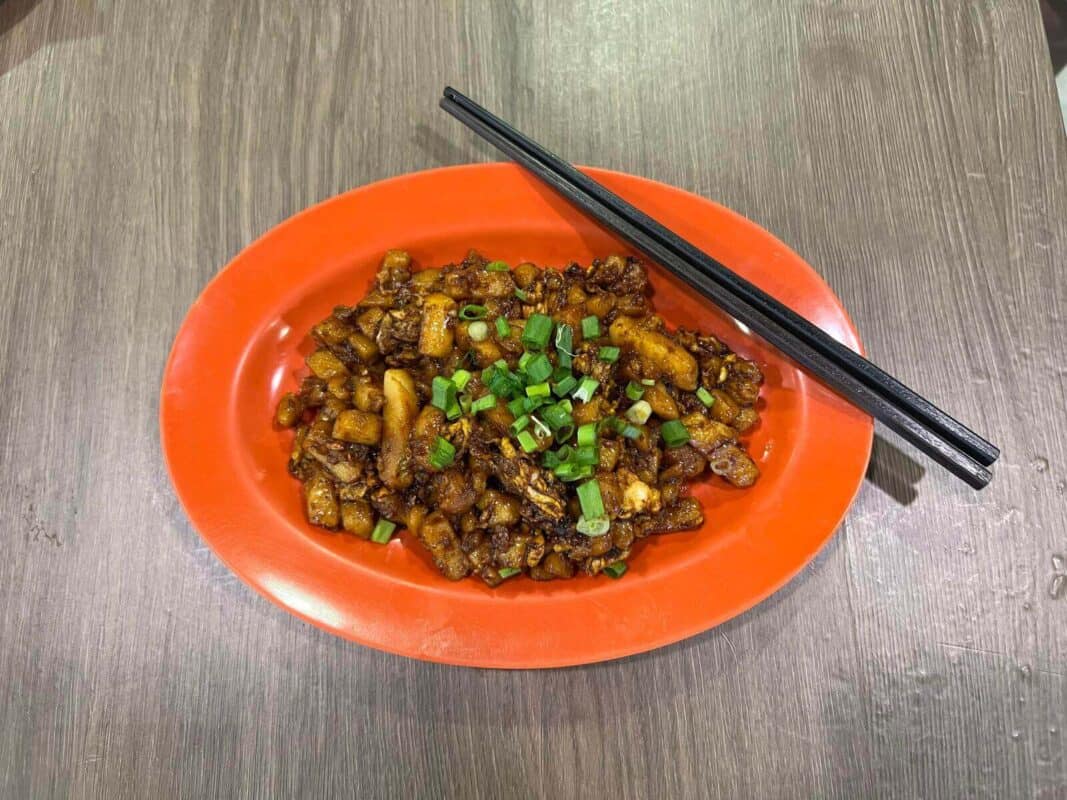 Singapore hawker food - A dish of Singaporean black carrot cake topped with chopped scallions, served on a orange plate with a pair of chopsticks resting beside it. The carrot cake pieces are stir-fried to a dark caramel color, offering a savory appearance on a textured gray table surface.