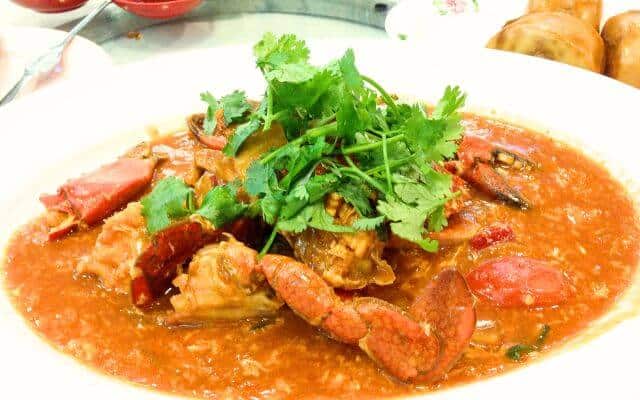 A dish of vibrant Singaporean chili crab, covered in a thick, spicy tomato-based sauce and garnished with fresh Chinese parsley. The succulent crab pieces are surrounded by a sauce that glimmers with hints of chili and garlic, served with mantou buns visible in the background, ideal for dipping.