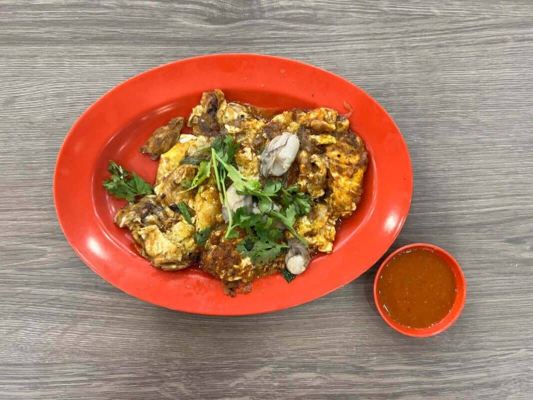 A Singaporean-style fried oyster omelette garnished with fresh Chinese parsley, served on a vibrant red plate alongside a small bowl of chili sauce. The omelette is golden brown, speckled with cooked oysters, and presented on a textured gray tabletop. It is one of the best hawker foods in Singapore.