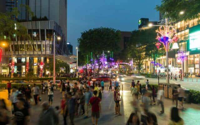 Orchard Road in Singapore bustling with people during the Christmas festive season, adorned with colorful lights and decorations. The scene features busy sidewalks, lit-up trees, and vibrant storefronts in the evening.