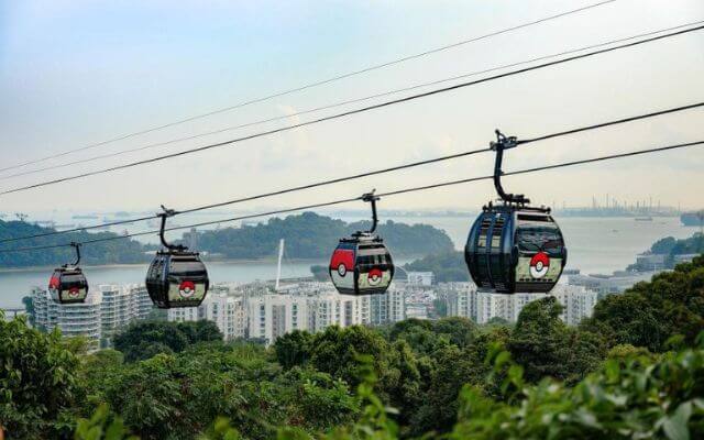Colorful cable cars decorated with Pokémon designs travel between Singapore and Sentosa Island, offering a panoramic view of lush greenery, residential buildings, and the distant coastline over the water.