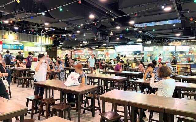 A food court in Singapore during an off-peak period, with diners seated at wooden tables under a ceiling adorned with string lights. Several food stalls are visible in the background, offering a variety of local dishes.