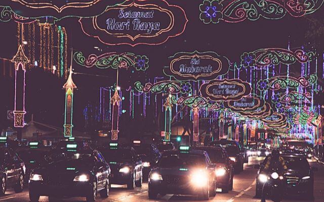 Street view of Selamat Hari Raya decorations in Singapore, with colorful and illuminated signs and designs hanging above the road, as cars drive beneath the festive lights at night.