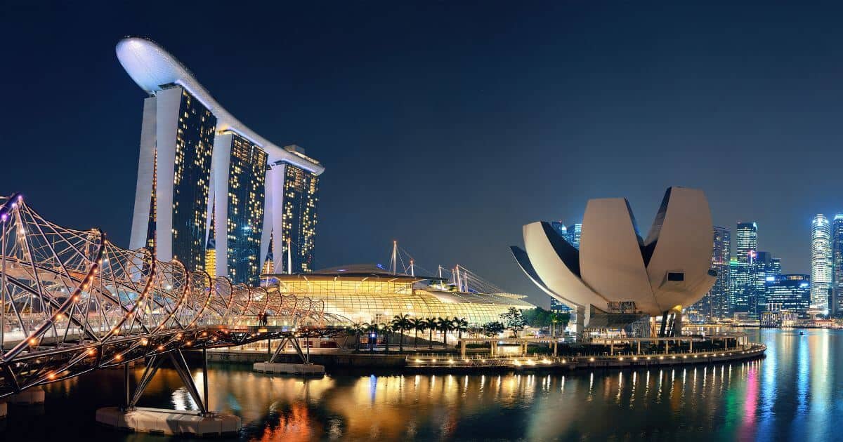 Night view of Marina Bay Sands Hotel and the ArtScience Museum in Singapore, illuminated with lights reflecting on the water. The iconic Helix Bridge is visible in the foreground, connecting the structures with the city skyline in the background.