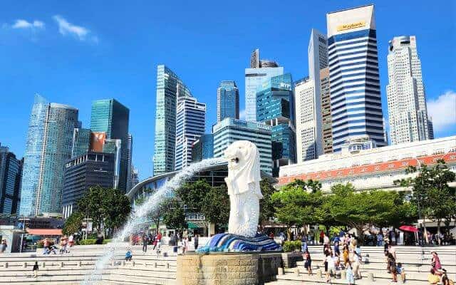 The iconic Merlion statue at Merlion Park in Singapore, spouting water into the bay with the city's financial district skyscrapers forming a striking backdrop. The park is lively with visitors enjoying the sunny day and scenic views.