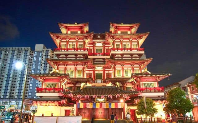 The Buddha Tooth Relic Temple in Singapore illuminated at night, showcasing its intricate traditional Chinese architecture with multiple tiered roofs and vibrant red and gold colors. The temple stands out against the night sky with a modern high-rise building in the background.