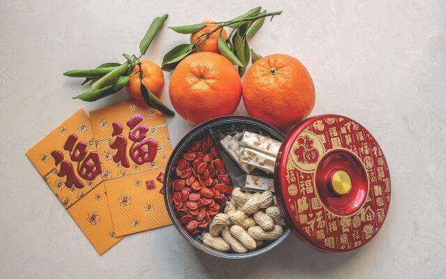 Traditional Chinese New Year items including mandarins, ang pao (red envelopes), and a round red box containing snacks like peanuts, candied fruits, and sweets. The arrangement is festive and symbolic of prosperity and good fortune.
