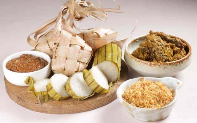 A traditional Hari Raya meal featuring ketupat (rice cakes wrapped in woven palm leaves), rendang (spicy meat stew), lemang (glutinous rice cooked in bamboo), and various sambals (spicy condiments) served on a wooden platter.