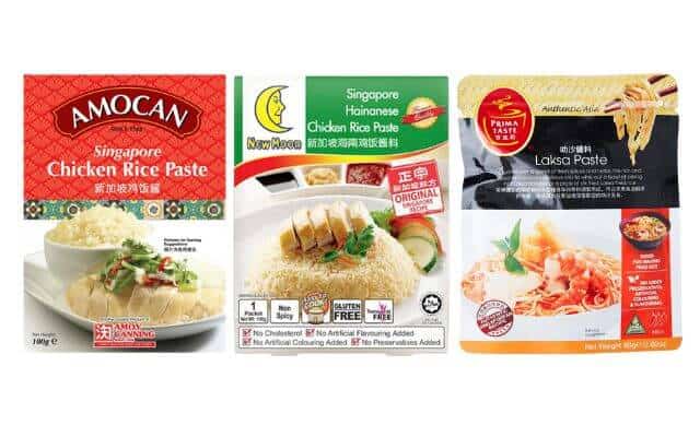 Showcases three different packaged Singaporean food pastes. From left to right, the first is "Amocan Singapore Chicken Rice Paste" in a red packet featuring a plate of chicken rice. The second is "New Moon Singapore Hainanese Chicken Rice Paste. The third is "Prima Taste Laksa Paste," presented in a package with an image of noodles in a rich laksa broth, garnished with shrimp and herbs.