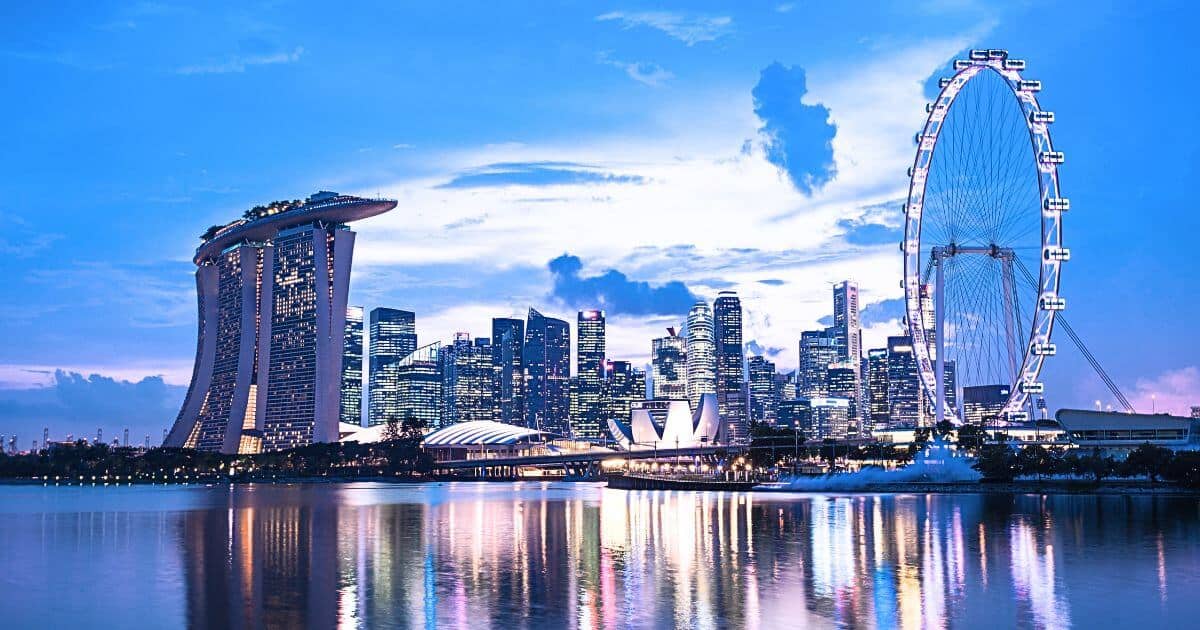 The iconic Singapore skyline at dusk. On the left is the Marina Bay Sands hotel, known for its boat-shaped rooftop, and to the right is the Singapore Flyer, a large observation wheel. The skyline is filled with illuminated skyscrapers reflecting on the calm waters below.