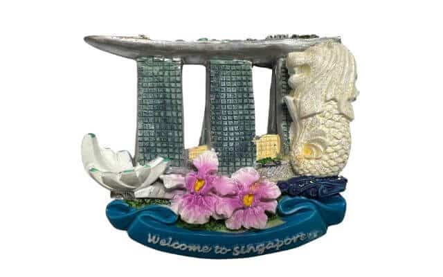 A decorative fridge magnet representing famous landmarks of Singapore. It features the Marina Bay Sands towers in the center, a large Merlion statue on the right, and pink flowers in the foreground. At the bottom, there is text that reads "Welcome to Singapore" on a blue banner. This magnet serves as a souvenir symbolizing iconic Singaporean attractions.