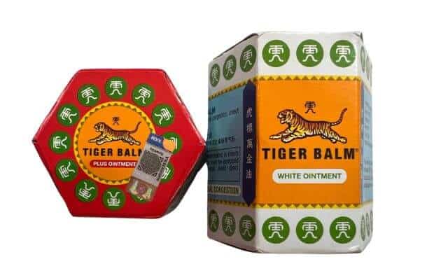 Two boxes of Tiger Balm ointment. The one on the left is in a red hexagonal container labeled "Tiger Balm Plus Ointment" with the iconic tiger logo and green Chinese characters around the edges. The box on the right is in a white and orange package labeled "Tiger Balm White Ointment." Tiger Balm is a popular remedy for pain relief and congestion.