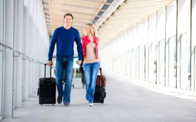 A couple walking through a brightly lit airport terminal, each pulling a wheeled suitcase on vacation.