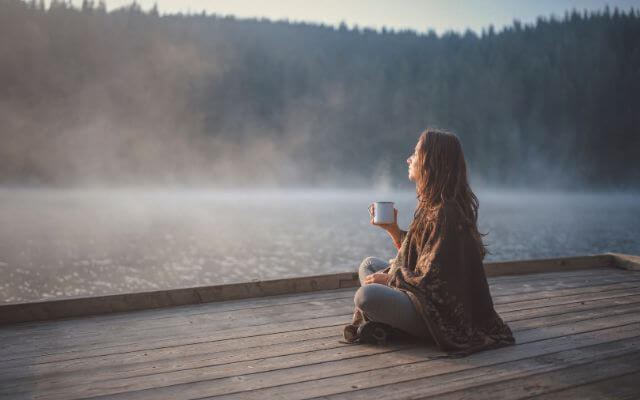 A woman relaxing on a wooden deck overlooking a misty lake, holding a cup of coffee.