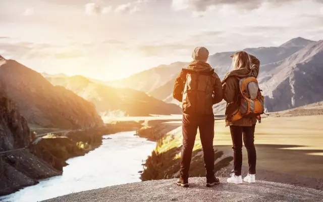 A couple stands on a rocky overlook, gazing at a river winding through a mountainous landscape at sunset. Dressed in outdoor gear with backpacks and sturdy trekking shoes, their footwear offers support and stability on rugged terrain, helping to prevent issues like plantar fasciitis during long hikes