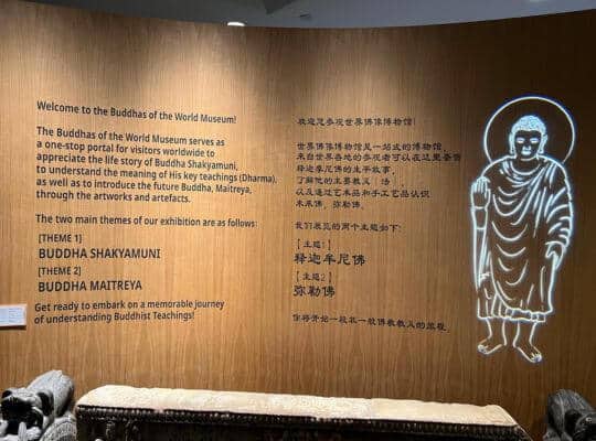 Interior signage of the Buddhas of the World Museum in Singapore. The text introduces the museum's focus on the teachings and stories of Buddha Shakyamuni and the future Buddha Maitreya, with the themes listed.