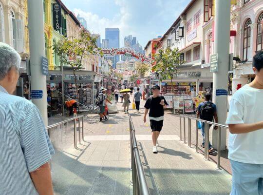 A bustling street market in Singapore's Chinatown, with colorful traditional shophouses lining the walkway. Stalls sell souvenirs, food, and other goods along the street.