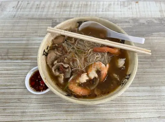 A bowl of prawn noodle soup featuring rice vermicelli, fresh prawns, slices of pork, and bean sprouts in a rich, flavorful broth, served with a side of sliced chili in soy sauce.