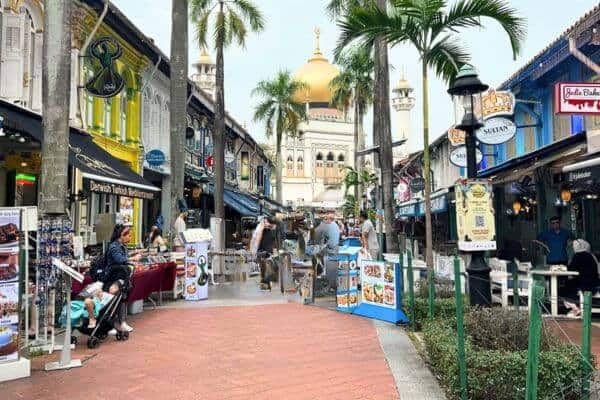 A vibrant street in Kampong Glam, Singapore, leads to the iconic Sultan Mosque with its golden dome and white minarets. The street is lined with colorful shophouses, home to restaurants, cafes, and stalls selling Middle Eastern and Malay-inspired goods.