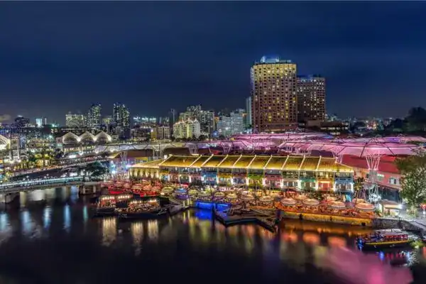 Night view of Clarke Quay in Singapore, with colorful lights illuminating riverside restaurants and bars under umbrella-shaped canopies. Reflections shimmer on the Singapore River as bumboats float by.